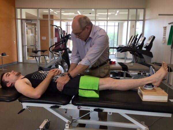 A man is being helped by a physical therapist who utilizes a microFET®2 in a gym.